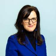 486086 Portrait headshot. The picture is taken in front of a blue background and the subject is wearing a sapphire blue blazer with 4 black buttons, and a black turtleneck shirt underneath. Her hair is dark brunette and her eyes are brown. She wears dark glasses, and blue dangling earrings. She is facing the camera, with shoulders slightly towards the right. 