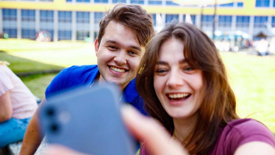 een meisje in een paars shirt en donker lang haar en een jongen in een blauw shirt met kort donker haar, lachen en maken een selfie met een smartphone.