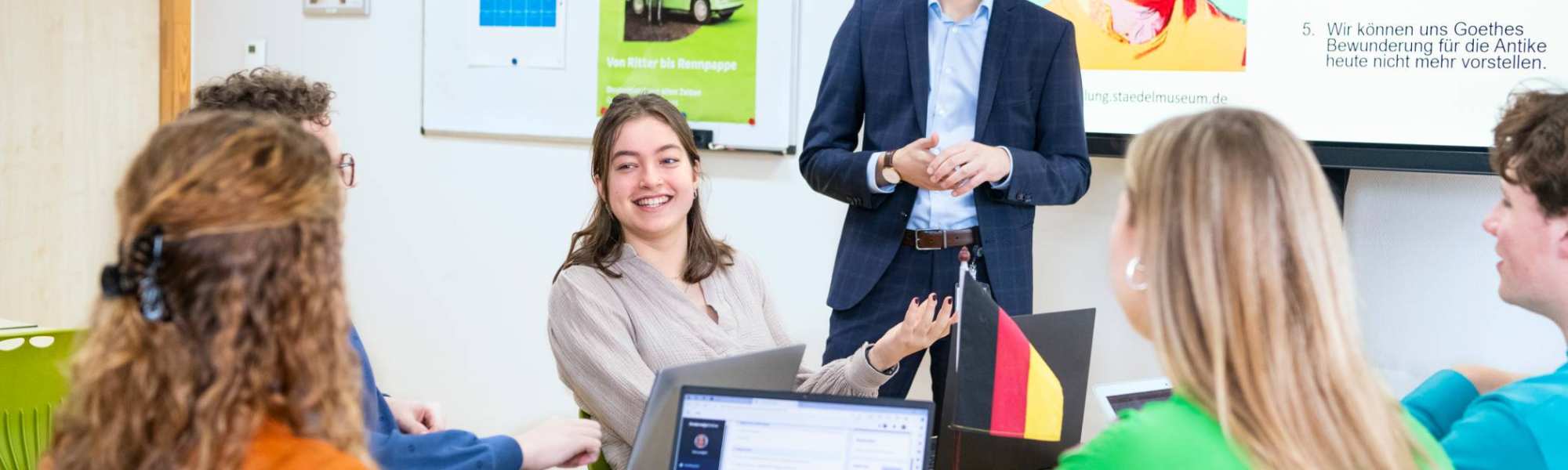 Studenten zitten samen aan tafels met laptops en boeken in het klaslokaal. De leraar staat ervoor en praat met hen mee.
