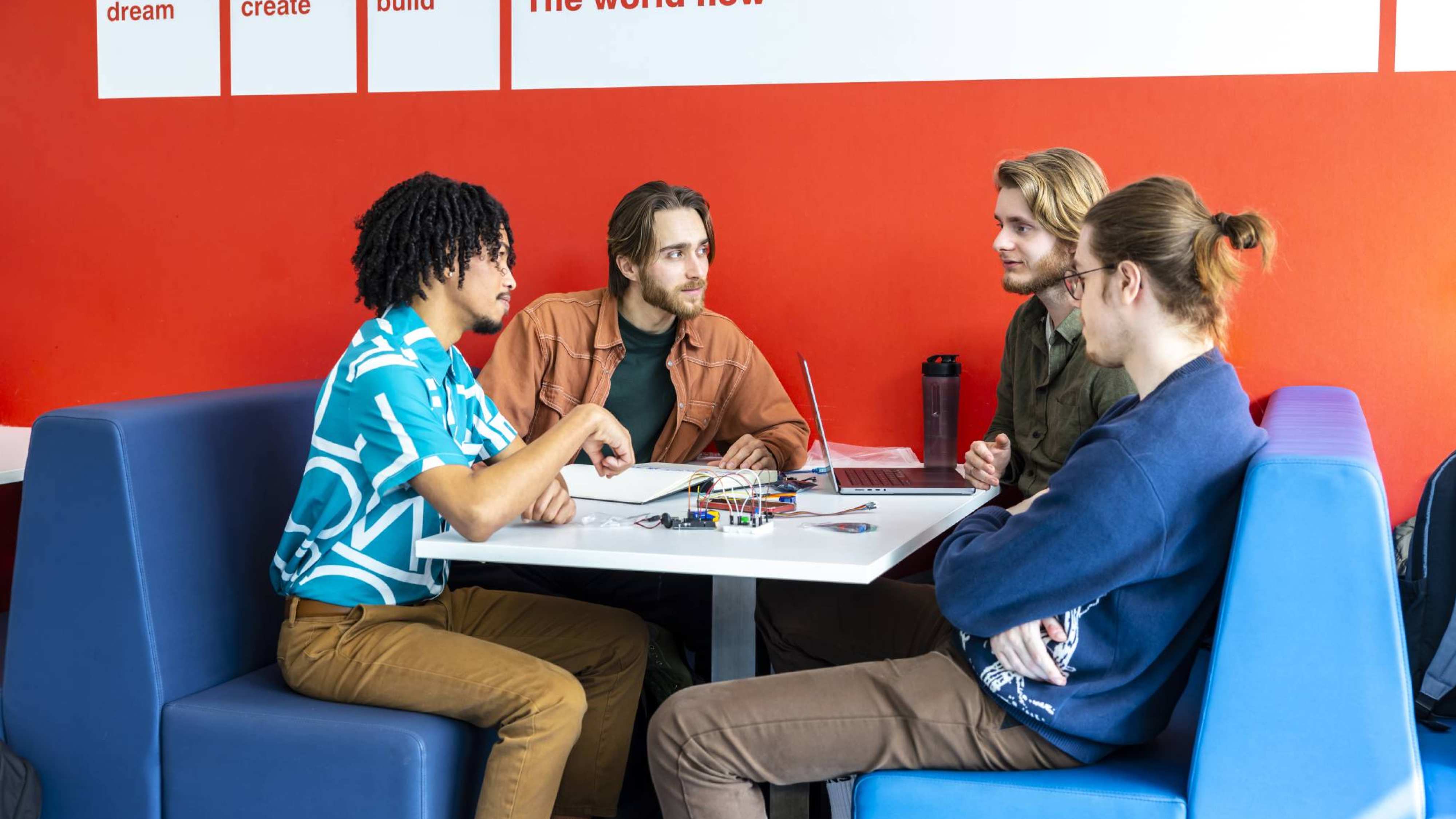 Studenten en docent in overleg met elkaar, bij blauwe banken en rode muur.